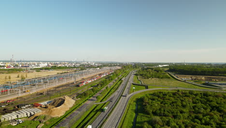 Aerial:-Driving-road-beside-green-forest-and-goods-station-in-Gdansk,Poland-during-sunny-day-with-blue-sky