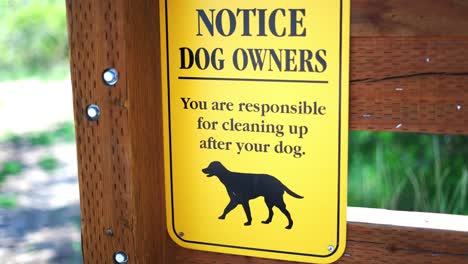 Notice-to-Dog-Owners-you-are-responsible-for-cleaning-up-after-your-pet-bright-yellow-sign-at-public-park-hiking-trail,-panning-back-and-forth-keeping-sign-in-frame,-in-4k-slow-motion