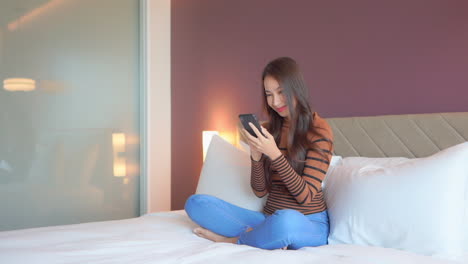 Pretty-Asian-Woman-Playing-Games-on-Mobile-Phone-while-Sitting-on-the-Made-up-Hotel-Bed-crossed-legs,-middle-shot