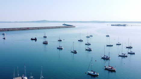 Sunny-Holyhead-harbour-breakwater-maritime-yachts-docked-along-transparent-calm-blue-shoreline-aerial-view-orbit-right