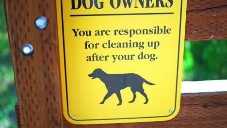 Notice-to-Dog-Owners-you-are-responsible-for-cleaning-up-after-your-pet-bright-yellow-sign-at-public-national-park-hiking-trail,-panning-from-top-to-bottom,-in-4k-slow-motion