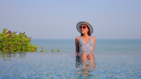 Stunning-girl-sitting-leaning-on-elbows-in-shallow-water-inside-infinity-pool-enjoying-gorgeous-tropical-scenery,-daytime-slow-motion