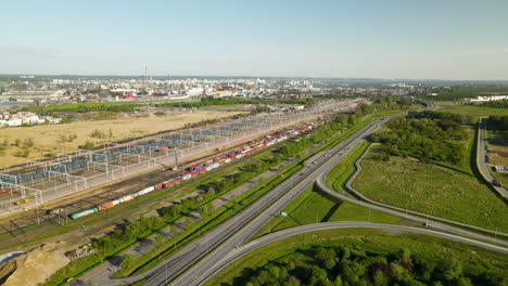 Aerial-View-of-a-Freight-Cargo-Train-Moves-on-Railway-out-from-Gdansk-Cargo-Terminal,-Cars-traffic-on-the-expressway-in-summer,-Gdansk-city-panorama