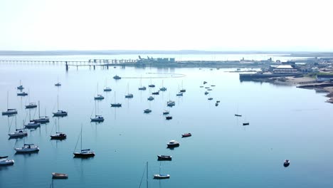 Sunny-Holyhead-harbour-breakwater-maritime-yachts-docked-along-transparent-calm-blue-shoreline-aerial-flyover-view