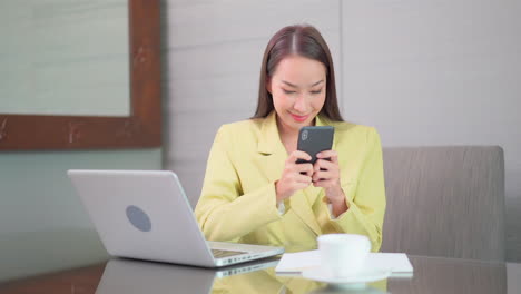 Asiatische-Geschäftsfrau,-Die-Auf-Dem-Bildschirm-Des-Handys-Tippt,-Während-Sie-Am-Schreibtisch-Vor-Dem-Laptop-Sitzt