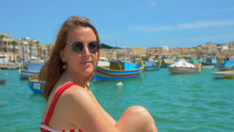 Chica-Europea-Con-Gafas-De-Sol-Se-Sienta-En-El-Puerto-Junto-Al-Luzzu-En-Verano-En-Marsaxlook,-Malta