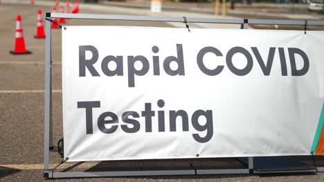 Rapid-Covid-19-Testing-site-sign-posted-outside-on-sunny-summer-day-in-empty-parking-lot-with-orange-cones-setup,-and-cars-driving-by-in-background