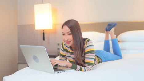 -Slow-motion-of-relaxed-Asian-woman-using-her-laptop-computer-lying-on-the-bed-with-her-legs-raised-at-hotel-room-wearing-casual-clothes,-working-from-home