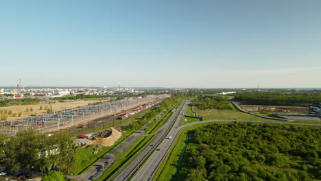 Aerial-View-of-the-Cars-Driving-Along-Highway-Road-Located-Near-Large-Train-Depot-With-Many-Cargo-Trains-in-Gdansk,-Poland,-summer-bird's-eye