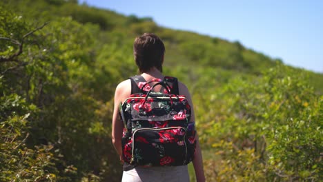 Women-girl-lady-with-short-hair-and-tank-top-on,-walking-or-hiking-lush-green-public-park-trail-outside-with-rose-covered-backpack-on-a-bright-sunny-hot-day-during-summer-in-2021