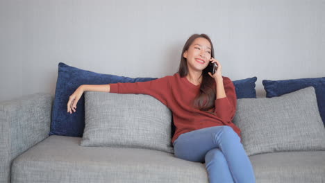 Beautiful-smiling-woman-talking-on-cell-phone,-Smiling-Asian-Thai-girl-sitting-on-the-couch-in-casual-clothes-and-having-mobile-phone-conversation-holding-smartphone-near-ear