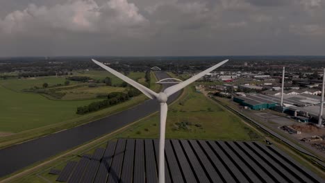 Aerial-panning-sideways-showing-a-wind-turbine-with-solar-panels,-biofuel-and-water-recycling-facility-in-Dutch-waterway-infrastructure-landscape