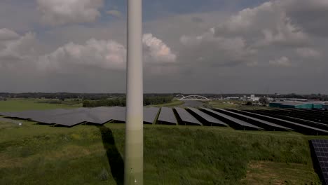 Aerial-ascend-revealing-a-rotating-wind-turbine-and-solar-panels-in-The-Netherlands-part-of-sustainable-industry-in-Dutch-flat-river-landscape-against-blue-sky-with-clouds