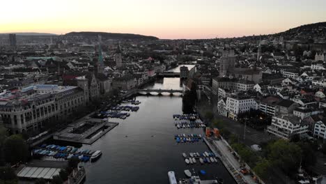 Aerial-flyover-over-Limmat-river-in-Zurich,-Switzerland-at-sunset-with-views-of-Grossmünster,-Frauenkirche,-St