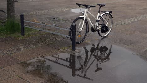 Bicycle-next-to-a-bike-stand-with-its-reflection-in-a-puddle-of-rain,-wide-shot