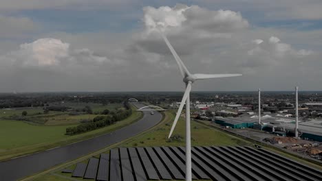 Approach-and-aerial-descend-showing-a-field-of-solar-panels-beneath-a-three-bladed-electricity-producing-wind-turbine-against-a-blue-sky-with-clouds