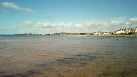 Elie-beach-and-harbour-viewed-from-the-beach-cafe-with-children-paddleboarding,-sun-shinning-and-beautiful-blue-sky