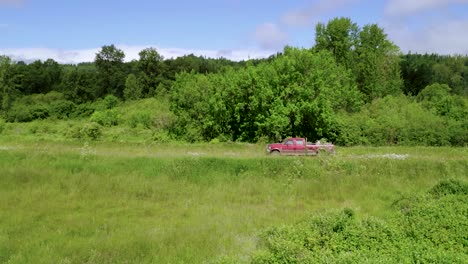 Red-Vehicle-Drives-On-The-Middle-Of-Green-Field-Beside-The-Lush-Forest