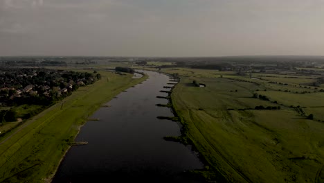 Ascending-backwards-aerial-movement-showing-IJssel-valley-with-meandering-river-and-floodplains-on-one-side-and-residential-area-on-the-other-side-in-The-Netherlands-on-an-overcast-day-during-sunset
