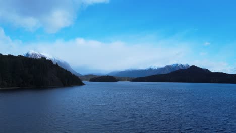 Lake-Nahuel-Huapi-on-a-sunny-day-between-clouds,-pronouncing-winter-with-its-white-topped-mountains-and-a-light-wind-bringing-small-waves-to-its-darkened-lake