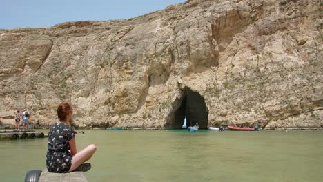 Frau-Im-Kleid-Sitzt-Auf-Felsen-Und-Beobachtet-Das-Binnenmeer-Und-Die-Boote,-Die-An-Sonnigen-Sommertagen-Durch-Die-Höhle-Auf-Der-Insel-Gozo-In-Malta-Fahren