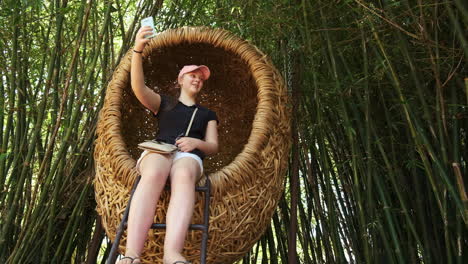 Young-girl-taking-selfie-in-wicker-egg-shaped-seat-in-oriental-bamboo-forest