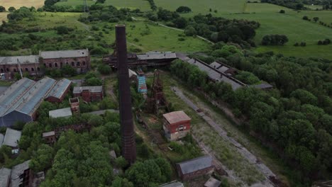 Abandoned-old-overgrown-coal-mine-industrial-museum-buildings-aerial-view-rising-above-chimney