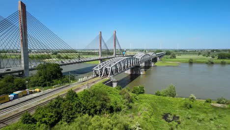 Aerial-view-of-dutch-train-crossing-a-railway-bridge-on-a-bright-sunny-day-surrounded-by-green-fields-and-river