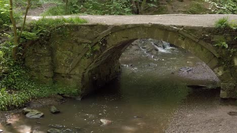 Peaceful-old-forest-stone-bridge-over-lush-hiking-woodland-trails-stream-slow-right