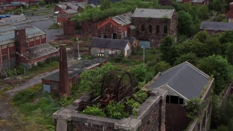 Abandoned-old-overgrown-coal-shaft-rusting-wheel-industrial-museum-buildings-aerial-rising-view
