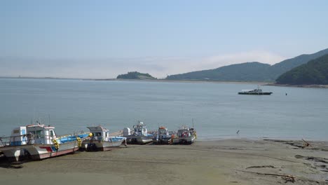 Fisher's-boats-stuck-in-the-mud-on-low-tide-at-Ganghwado-island-in-South-Korea,-One-shio-is-floating-in-sea-water,-haze-over-mountain-peaks,-Red-navigational-buoy-floating-in-Yellow-sea