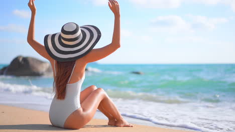 A-young-woman-in-a-bathing-suit-and-floppy-sun-hat-sits-on-a-sandy-beach-raising-her-arms-in-welcome-to-the-incoming-surf