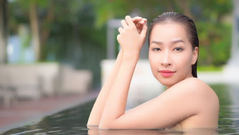 Beautiful-Asian-woman-with-wet-hair-bathing-in-a-pool-looking-at-camera-while-leaning-elbows-on-the-edge-of-the-swimming-pool