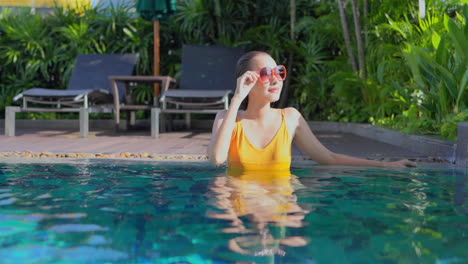 American-Woman-Inside-Swimming-Pool-Water-Leaning-on-the-Side-of-the-Pool-and-Touching-Sunglasses'-frame-in-the-tropical-Hotel-lounge-in-Miami,-static-slow-motion