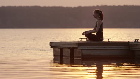 Young-woman-meditating-in-lotus-position-on-jetty-at-lake-at-sunset