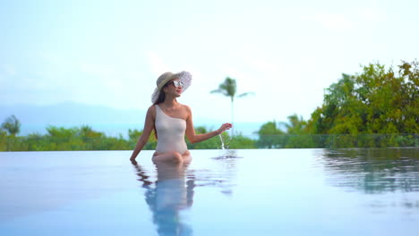Asian-lady-in-white-monokini-and-hat-sitting-on-infinity-pool-border-on-lush-tropical-greenery-background-in-luxury-Hotel-in-Bali-on-sunset