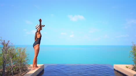 Woman-in-black-bathing-suit-and-striped-sunhat-raises-arms-standing-on-stony-edge-of-the-rooftop-infinity-pool-enjoying-stunning-seascape