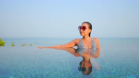 Portrait-shot-of-Thai-woman-in-sunglasses-resting-inside-infinity-pool-leaning-on-the-arm-on-the-edge-of-pool,-face-reflection-in-water-surface-daytime,-endless-sea-on-background