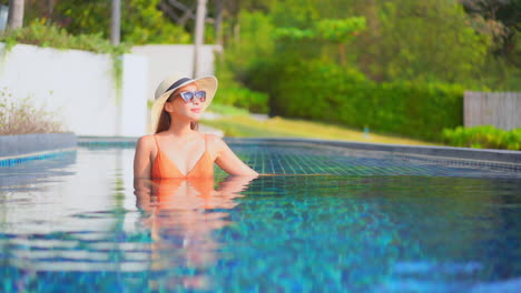 Portrait-of-attractive-Asian-woman-relaxing-inside-tropical-hotel-swimming-pool-water-wearing-orange-swimsuit,-sunglasses-and-beach-hat-in-Phuket-Thailand,-bust-shot-slow-motion