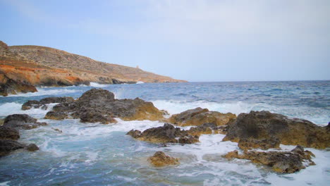 Massive-waves-and-whitewash-smashing-into-beach,-rocks-and-cliffs-in-slow-motion-alongside-rocky-coast-of-Ghar-Lapsi,-Malta