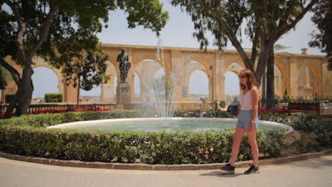 Joven-Mujer-Con-Estilo-Caminando-A-Lo-Largo-De-La-Fuente-De-Manantial-De-Agua-Mientras-El-Agua-Salpica-En-Cámara-Lenta-En-Los-Jardines-De-La-Barraca-Superior-En-Valletta,-Malta