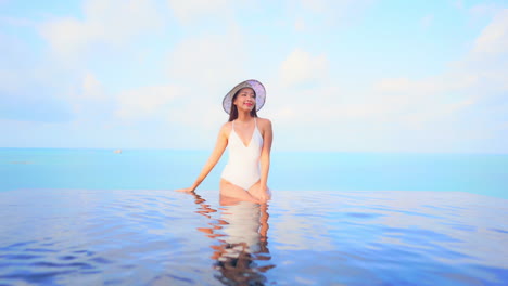 Eine-Atemberaubende-Frau-In-Einem-Weißen-Badeanzug-Sitzt-Am-Rand-Eines-Infinity-Pool-Ozeans-Im-Hintergrund