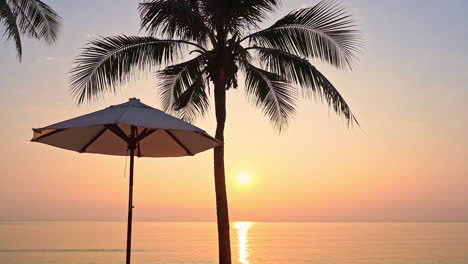 Perfect-red-sun-sunset-view-from-tropical-hotel-resort-lounge-near-the-sea,-beach-umbrellas,-and-coconut-palm-silhouette-in-the-foreground
