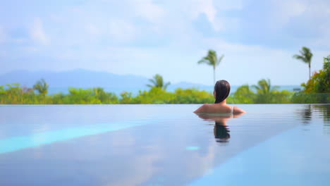 Back-view-of-woman-swimming-inside-pool-of-tropical-hotel-with-Palms-and-Mountains-panorama-on-background-in-Bali