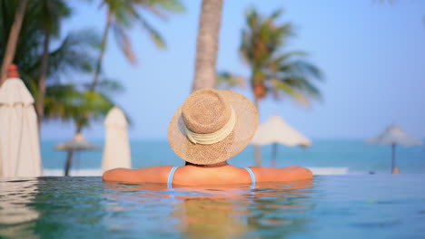 Back-View-of-the-Young-Woman-in-Infinity-Swimming-Pool-wearing-a-Straw-Hat-and-leaning-her-Arms-on-the-Edge-of-the-Pool-in-Tropical-Hotel-in-Malaysia