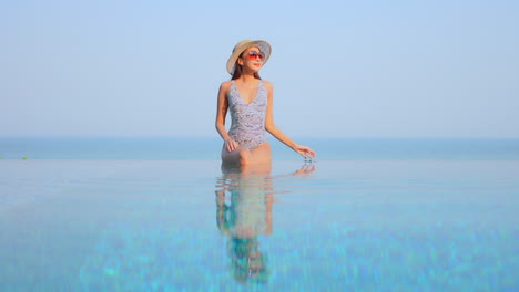 Pretty-woman-in-swimwear-sitting-on-infinity-pool-border,-touching-her-sunhat-and-raising-hands-up-with-a-stunning-view-of-tropical-sea-horizon-in-Phuket,-Thailand,-static-copy-space