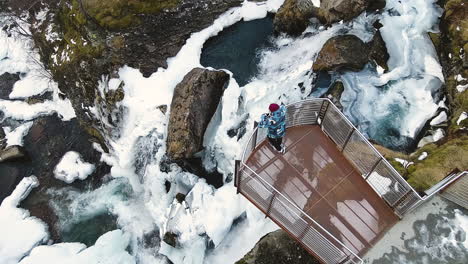 Man-On-Observation-Platform-In-Steep-Cliff-With-Overview-Of-Icy-Waterfall-In-Town-Of-Geiranger-In-Norway-During-Winter-Season