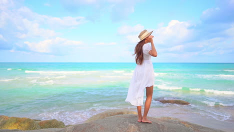 Lonely-sexy-exotic-female-in-a-white-dress-standing-on-rock-in-front-of-tropical-sea-waves-holding-floppy-summer-hat,-full-frame-slow-motion