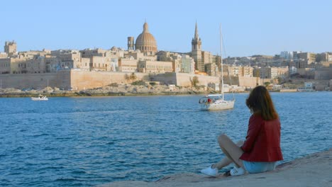 Eine-Frau,-Die-Am-Ufer-Sitzt-Und-Liest,-Mit-Der-Stadt-Valletta-Am-Horizont