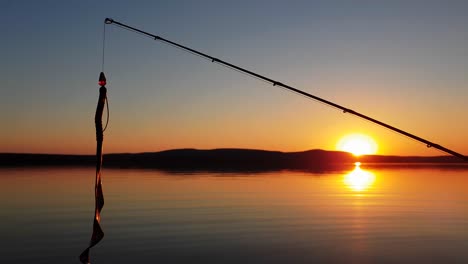 Ein-Barschfischköder,-Der-Von-Einer-Angelrute-Mit-Einem-Perfekten-Sonnenuntergang-über-Einem-Ruhigen-See-Im-Hintergrund-Baumelt